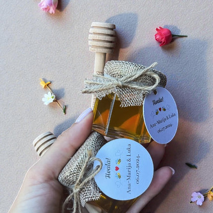 Small bottle of honey with a wooden dipper, tied with burlap ribbon, held by a hand on a light background with flowers.