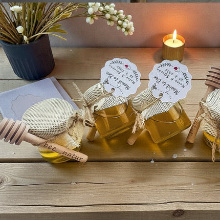 Three jars of honey with wooden spoons on a wooden surface with candles and flowers in the background.